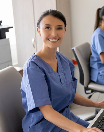 Woman in scrubs smiling working on a computer at a desk